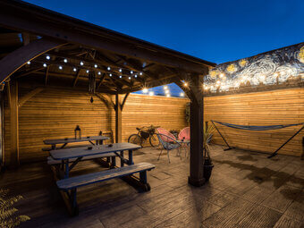 The outdoor patio at dusk, with a covered wooden gazebo, picnic benches, rattan egg chairs, a hammock, festoon lighting, and a starry-night mural on the fence