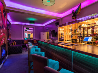 The Tipsy Unicorn Bar, a backlit bar on the ground floor with a mirrored bottle display, purple ambient lighting, and lounge seating