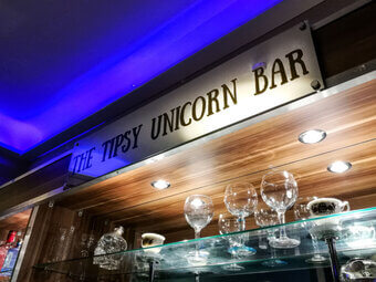 The illuminated Tipsy Unicorn Bar sign above a glass shelf of cocktail glasses, lit by blue ambient lighting